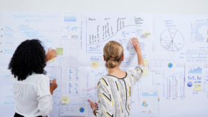 Dos professionals organitzant gràfics, mètriques i fases d'una campanya de màrqueting digital en un mural de treball