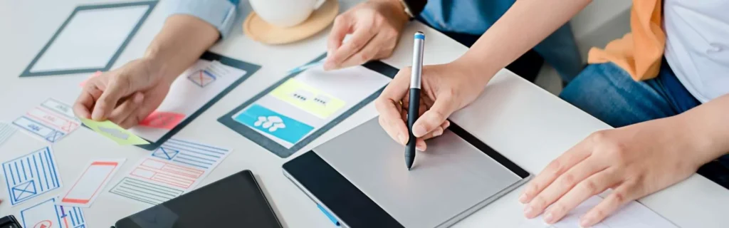 Primer plano de un equipo de diseñadores trabajando en el wireframe de una landing page sobre un escritorio, utilizando una tableta gráfica, notas adhesivas y bocetos de interfaz de usuario.