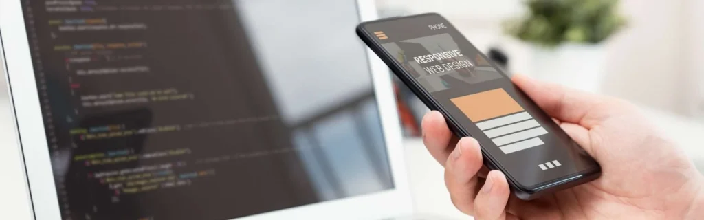 Mano sosteniendo un smartphone que muestra un diseño web responsive frente a una pantalla de ordenador con líneas de código de programación